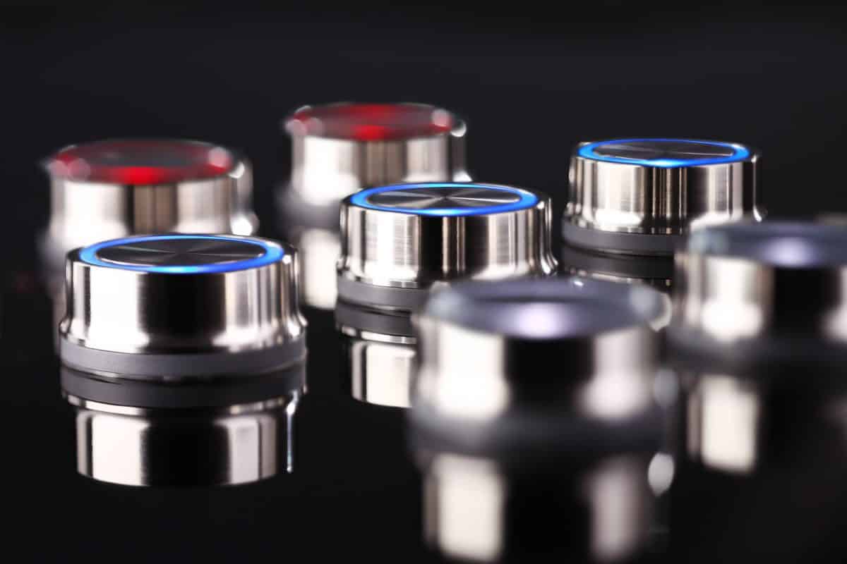 A group of RVA stainless steel round pushbuttons in soft focus, with their blue illumination rings contrasting against the black background.
